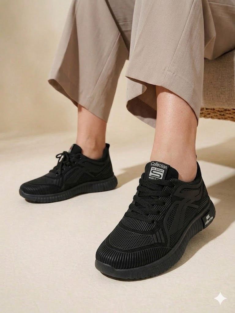A high-quality lifestyle product shot of a person sitting and wearing black "MK Collection" athletic sneakers. The shoes feature a textured, breathable mesh upper with a distinct wavy pattern on the sides and a chunky, ribbed black sole. The person is wearing light beige, wide-leg trousers that drape over the top of the sneakers. The lighting is soft and natural, emphasizing the matte black finish of the shoes against a neutral, minimalist background.