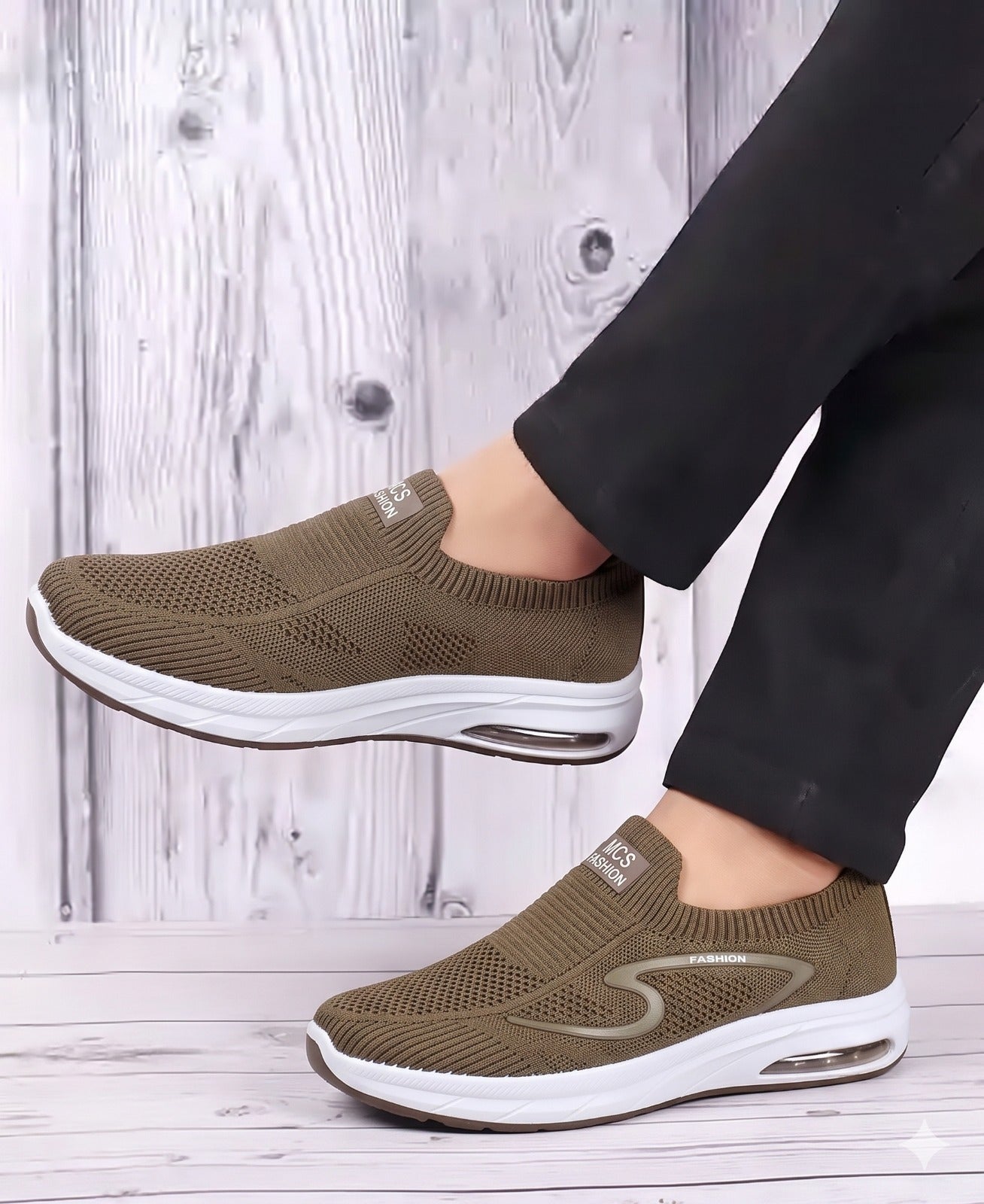Brown slip-on sneakers with white soles worn by a person on a wooden floor.