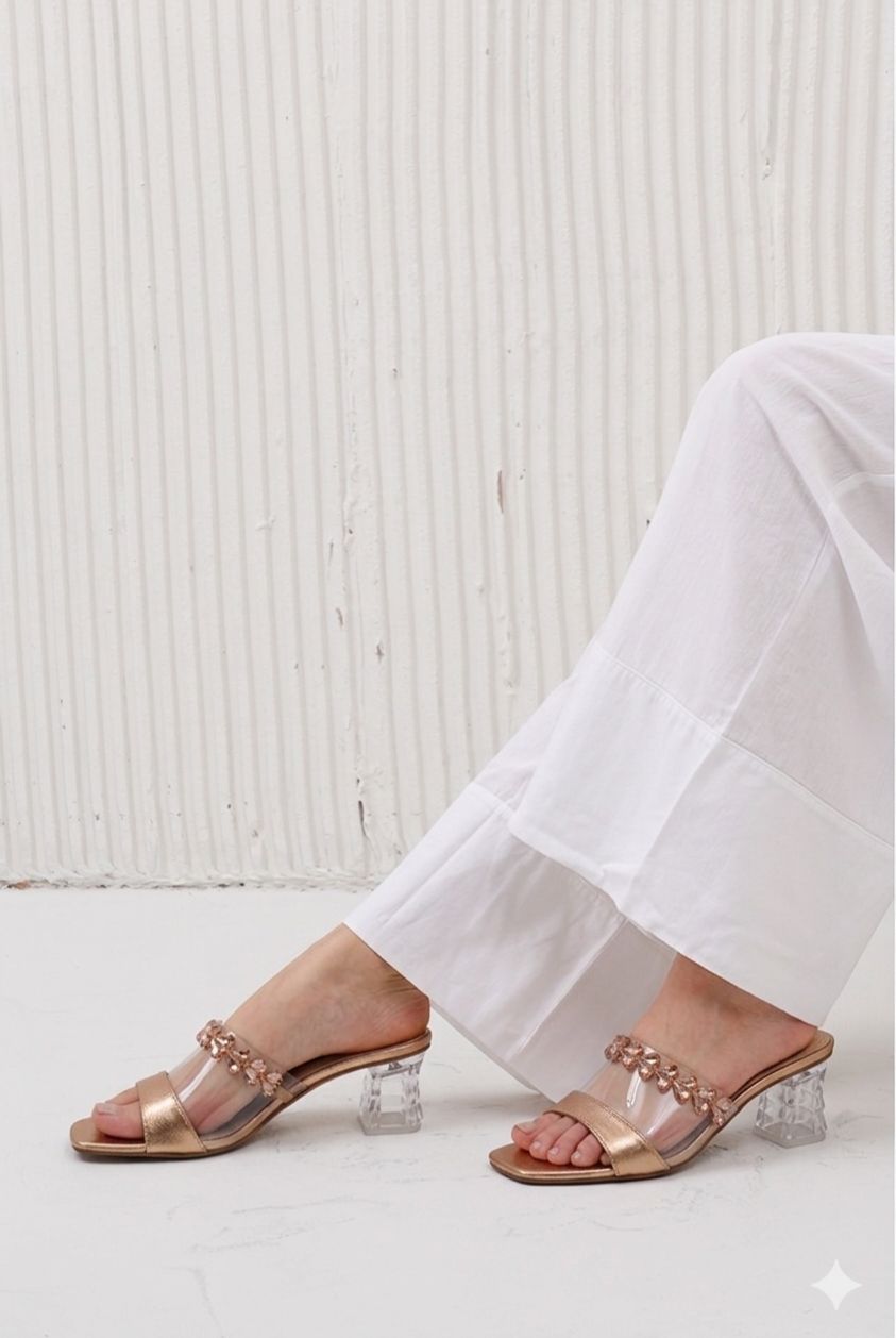 Gold sandals with clear heels worn by a person in white pants against a light background