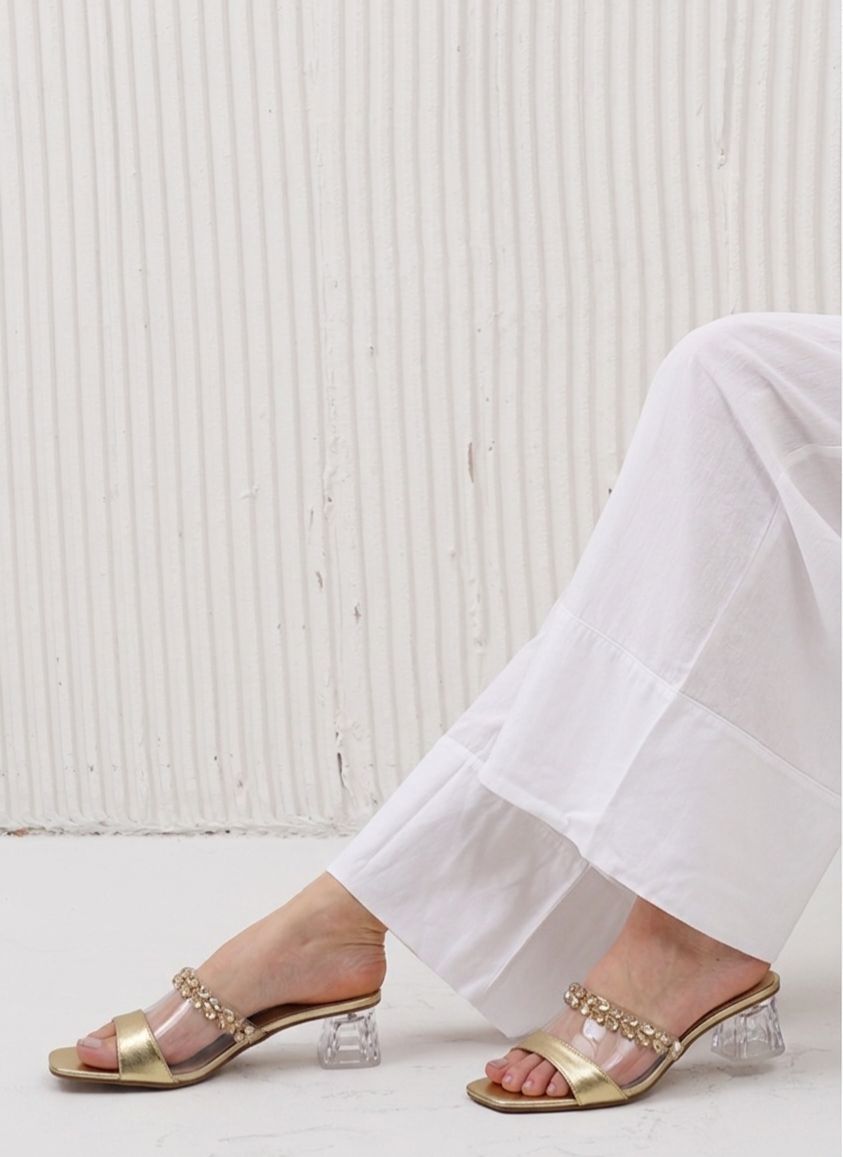 Gold sandals with crystal details worn with white pants against a light background