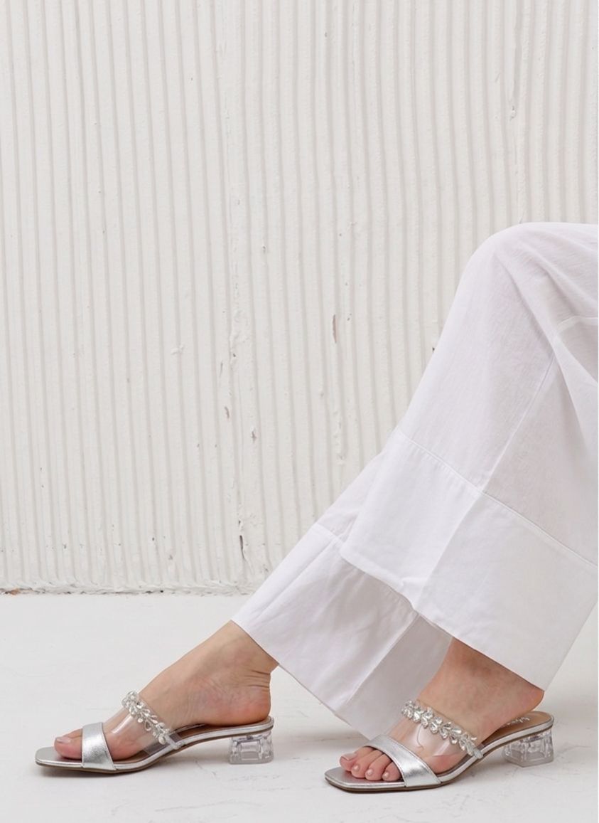 Silver sandals with clear straps worn with white pants against a light background