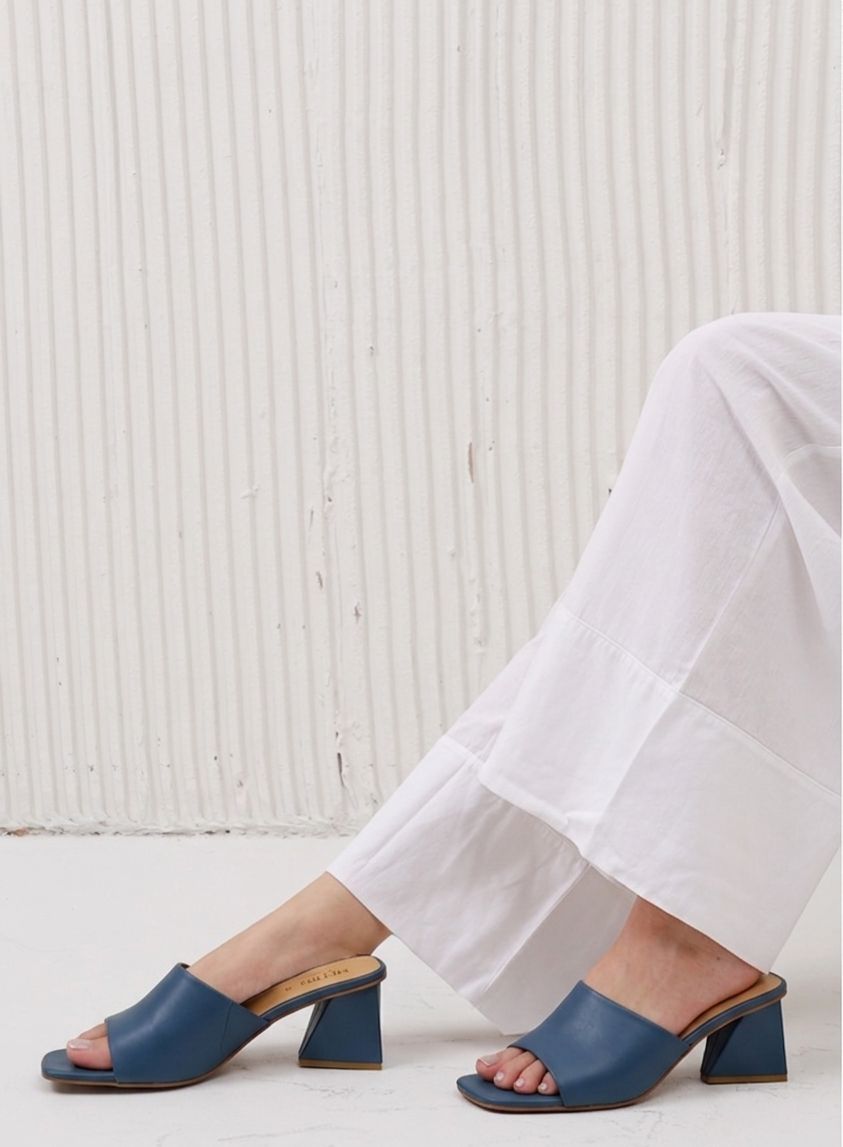 Blue sandals worn with white pants against a light wooden panel background