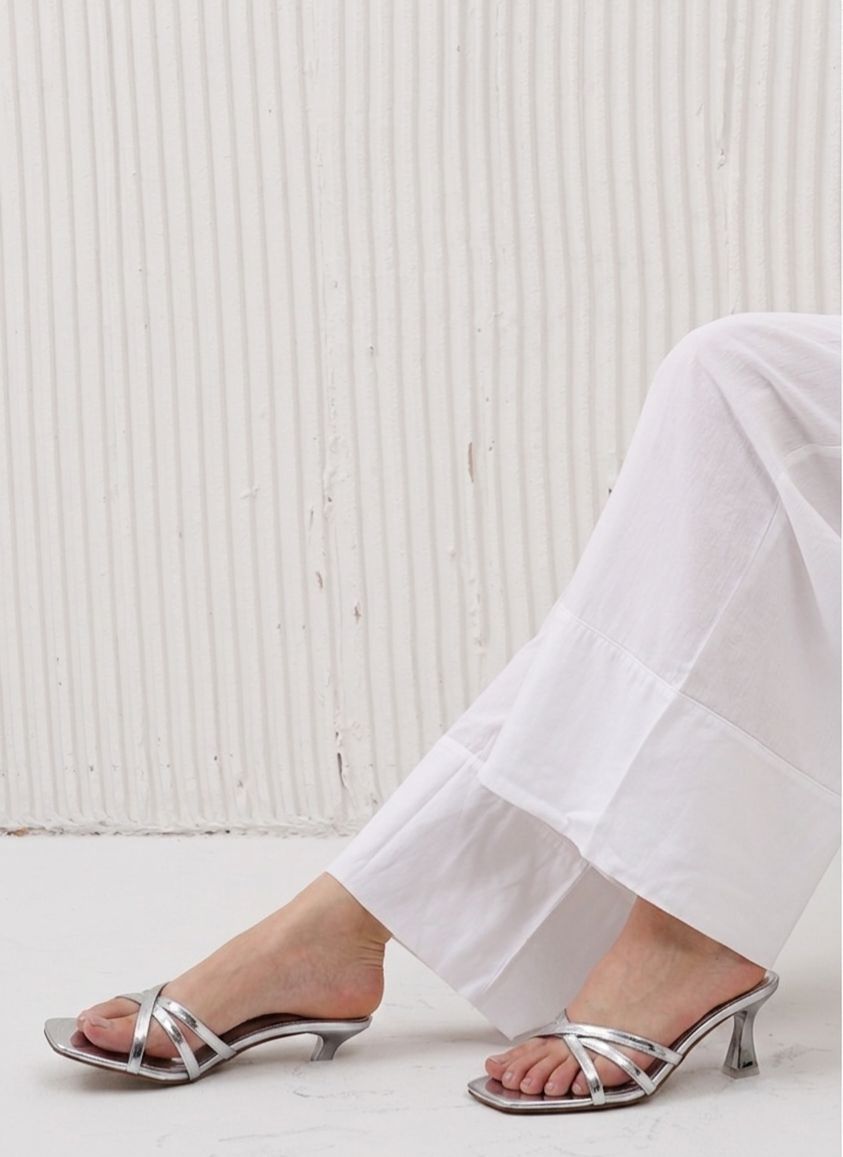 Silver high-heeled sandals worn with white pants against a light wooden background