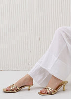 Gold high-heeled sandals worn with white pants against a white background