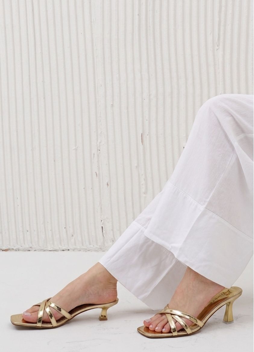 Gold high-heeled sandals worn with white pants against a white background