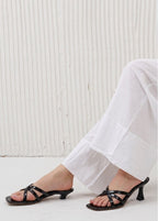 Person wearing black strappy high-heeled sandals with white pants against a light background
