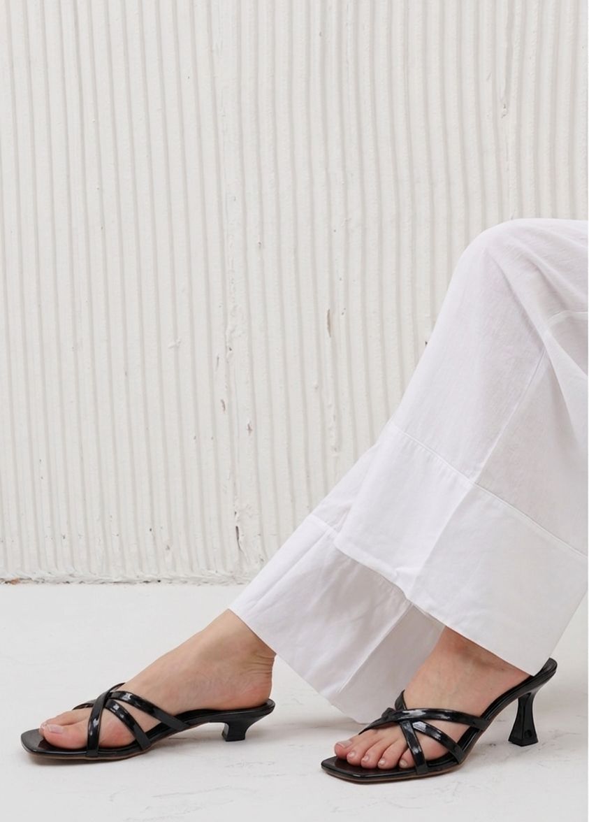 Person wearing black strappy high-heeled sandals with white pants against a light background