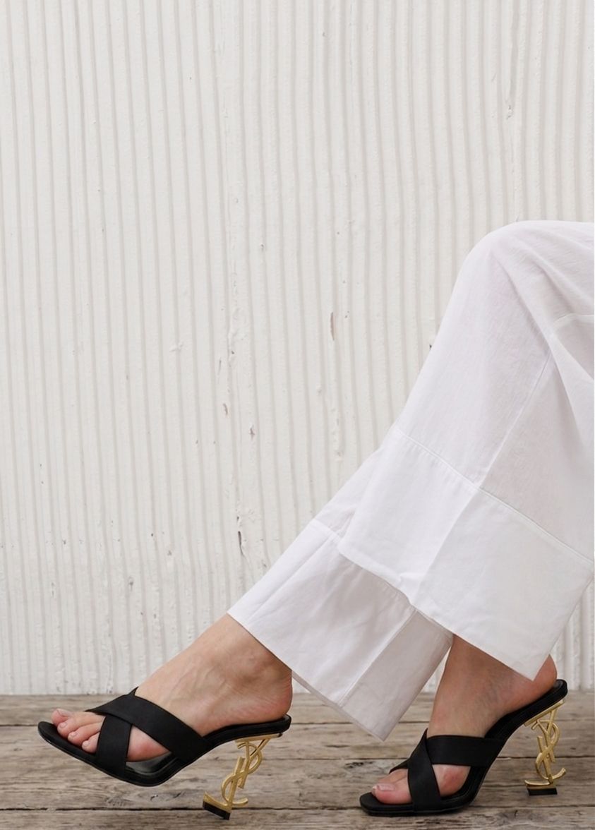 Black high-heeled sandals worn with white pants on a wooden floor.