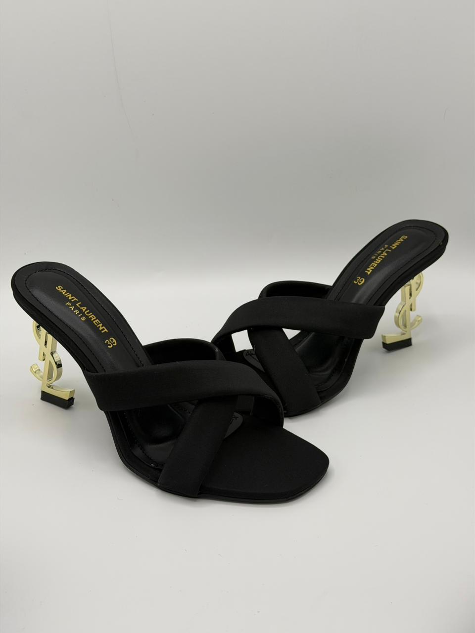 Black high-heeled sandals with gold accents on a light gray background
