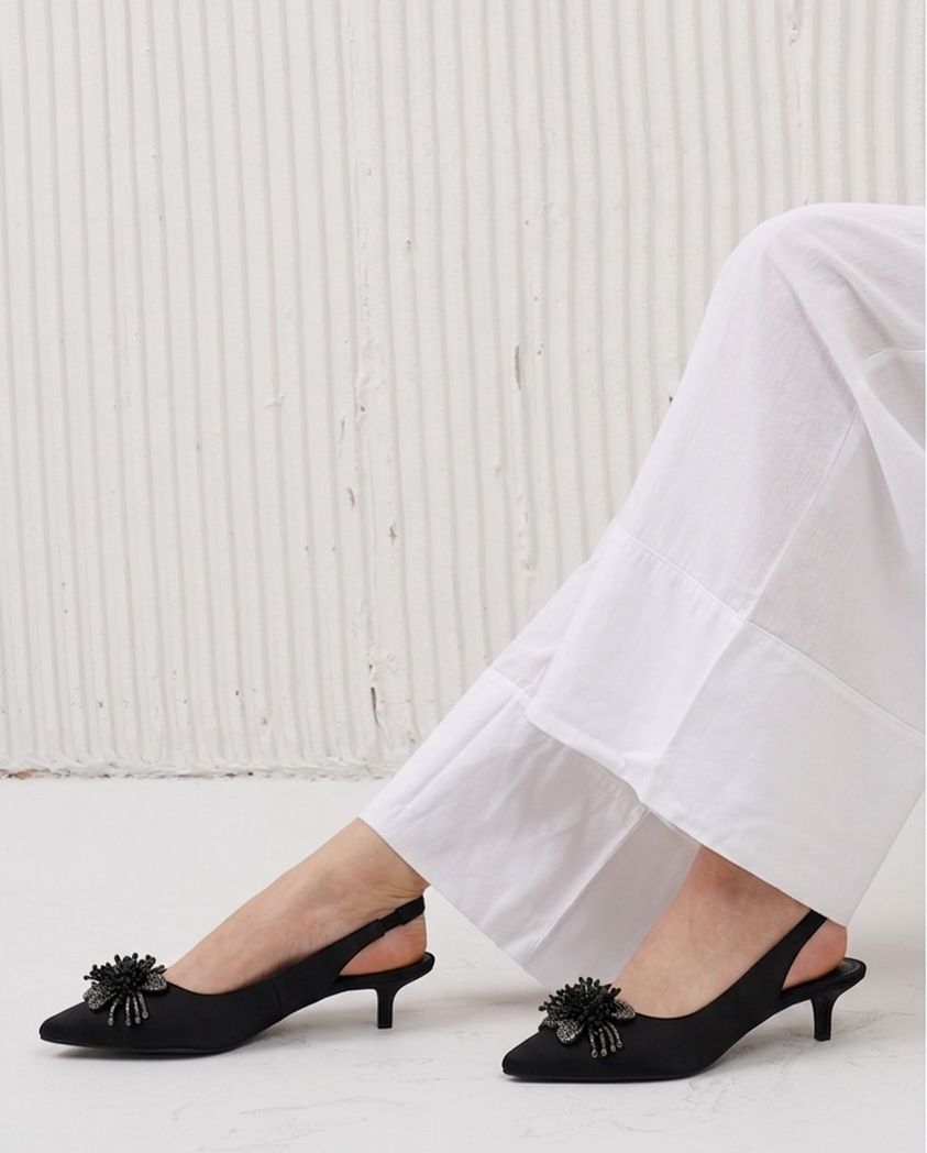 Black kitten heel shoes with floral details worn by a person in white pants against a light background