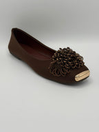 Brown flat shoe with decorative embellishment on a light gray background