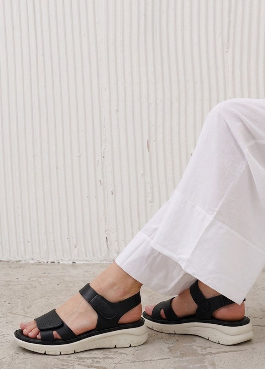 Person wearing black sandals with white soles on a light wooden floor.