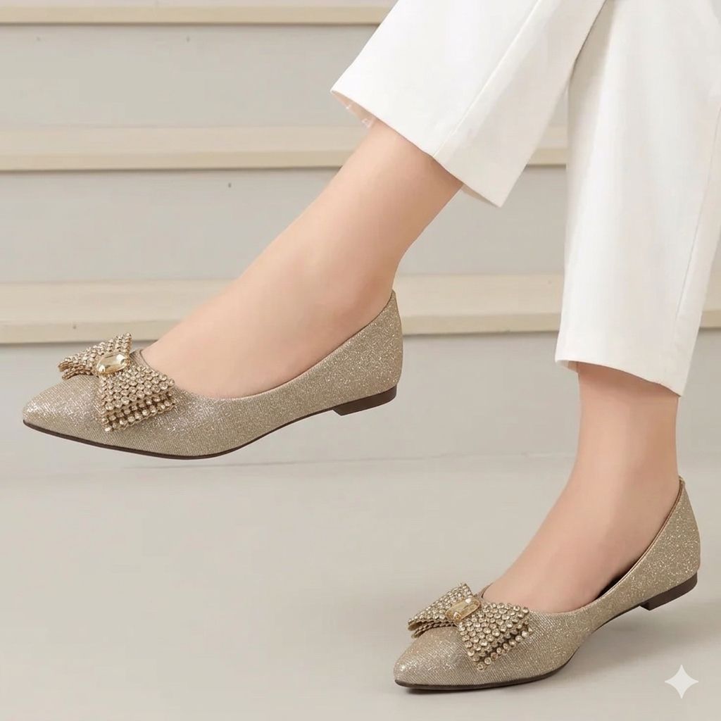 Gold glittery flats with embellishments worn by a person in white pants on a neutral background