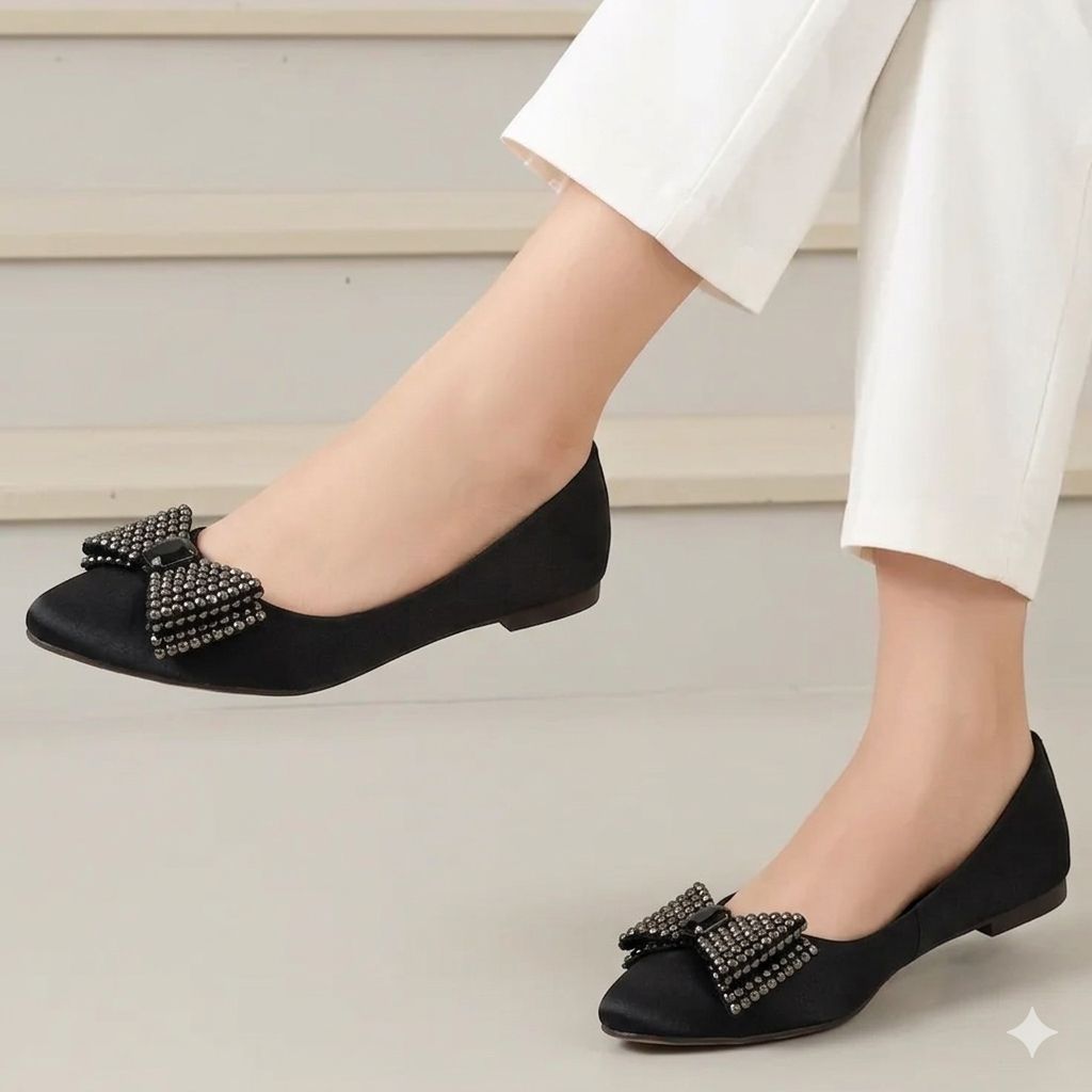 Black flat shoes with decorative bows worn by a person in white pants on a neutral background