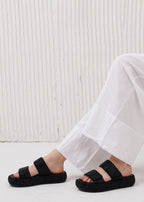 Person wearing black sandals with white pants against a light wooden panel background