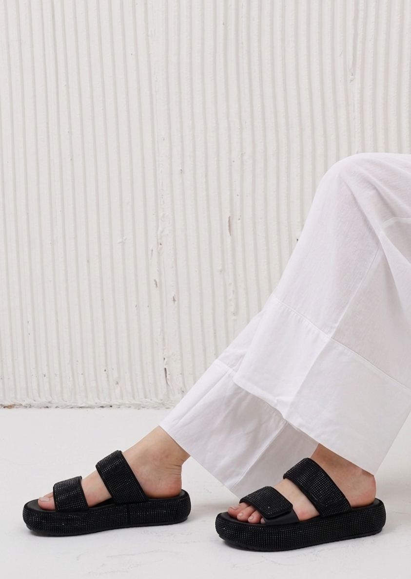 Person wearing black sandals with white pants against a light wooden panel background