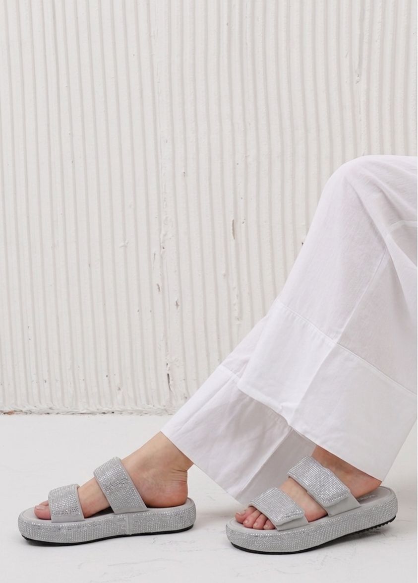 Gray sandals worn with white pants on a light wooden floor.