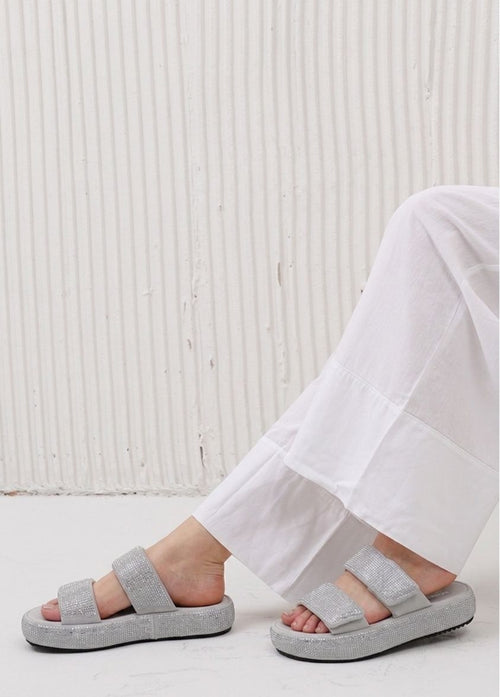 Gray sandals worn with white pants on a light wooden floor.