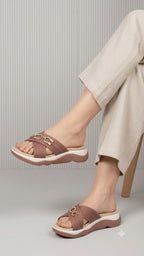 A person wearing taupe-brown slide sandals with a metallic gold horsebit detail on the straps, paired with light beige linen trousers, seated on a wooden chair against a gray ribbed background.