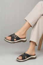 A person wearing black slide sandals with wide straps and a gold-tone horsebit detail, paired with light beige cropped pants, seated on a wooden chair against a gray ribbed background.