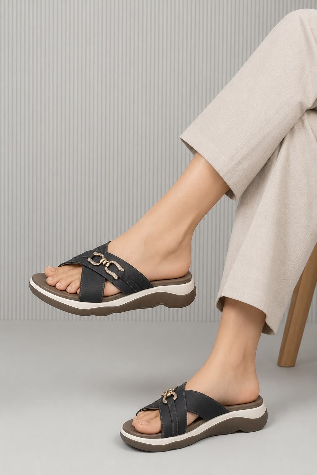 A person wearing black slide sandals with wide straps and a gold-tone horsebit detail, paired with light beige cropped pants, seated on a wooden chair against a gray ribbed background.