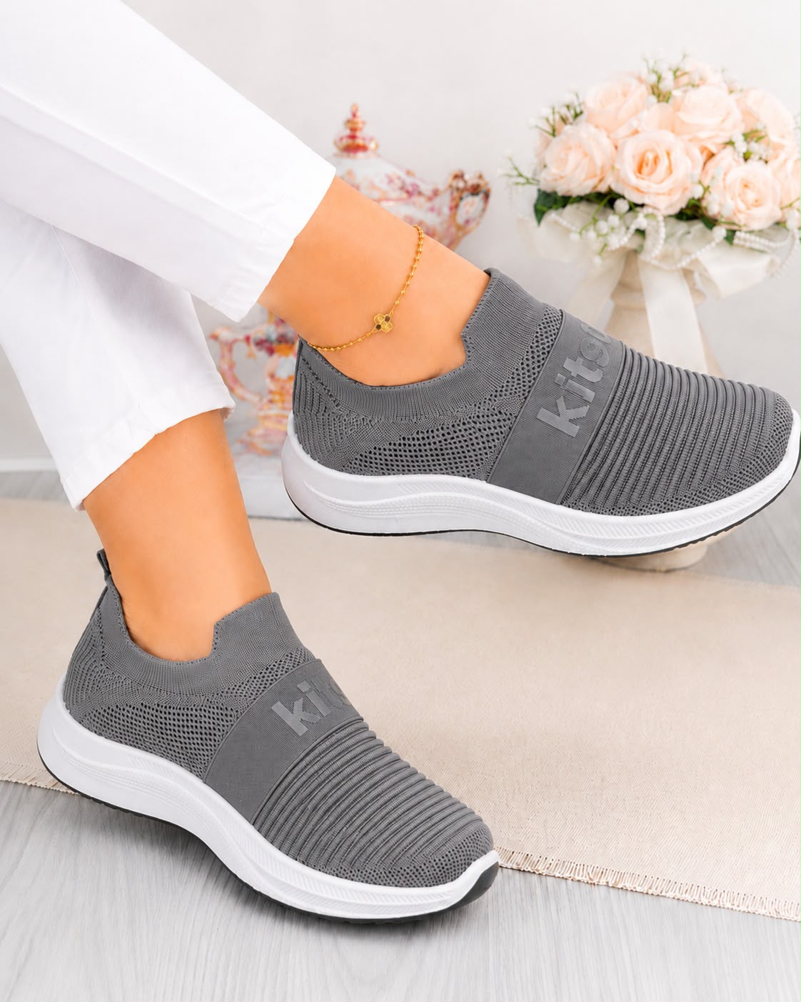 A pair of women's gray slip-on knit sneakers with white soles, shown being worn with white pants. The shoes feature a textured knit upper and a debossed 'kit' logo on the side.