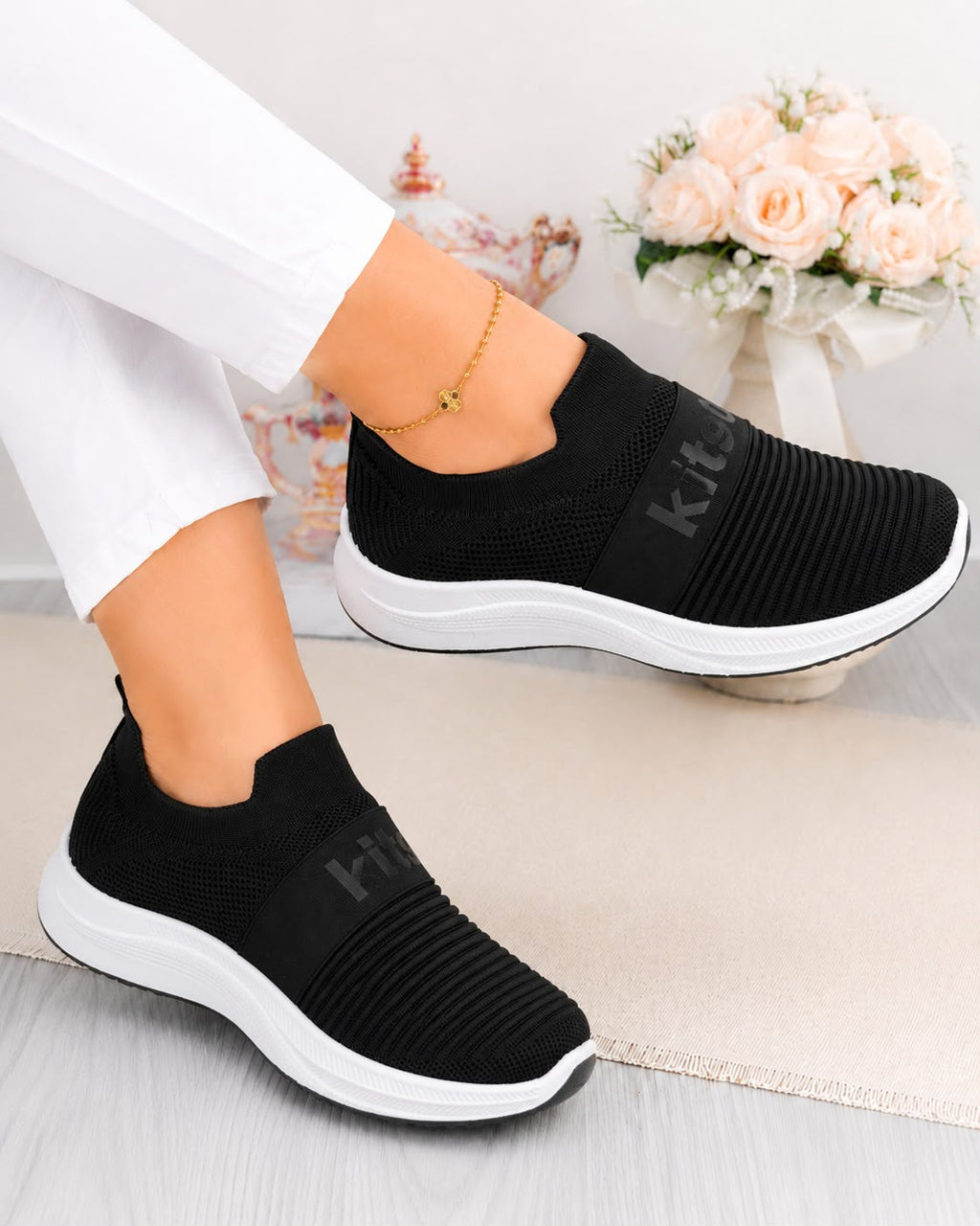 A pair of women's black slip-on knit sneakers with white soles, shown being worn with white pants. The shoes feature a ribbed knit upper and a dark gray 'kit' logo on the side.