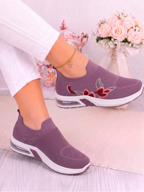 A pair of women's slip-on knit sneakers in a muted mauve-purple color, featuring a red and green floral embroidery on the side and a white sole with a clear air cushion. The shoes are modeled with white pants, shown in a styled indoor setting with a pink rug and flowers in the background.
