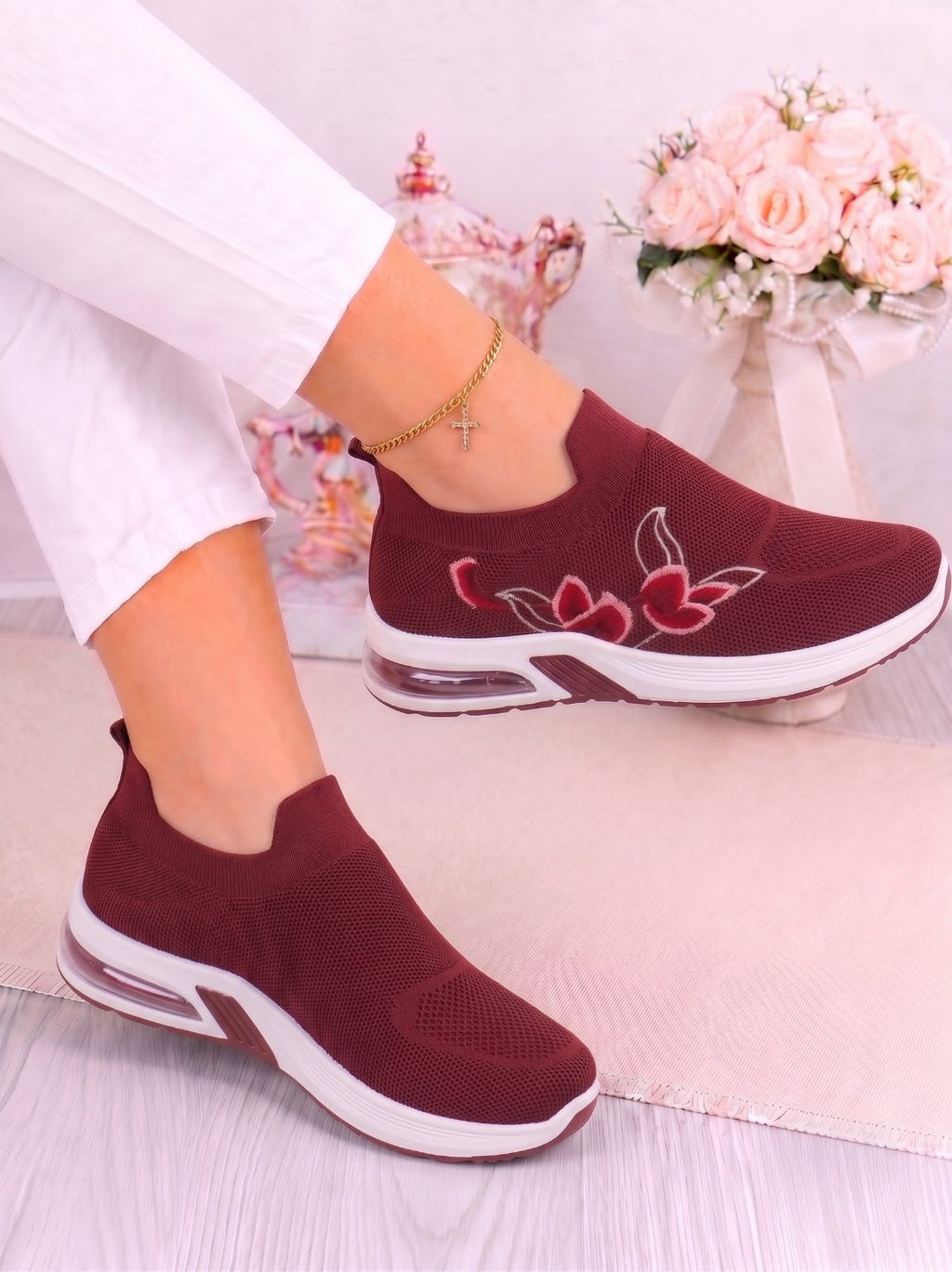 A pair of women's slip-on sneakers in burgundy with white soles and red floral embroidery on the side, shown being worn with white pants.