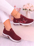 A pair of women's slip-on sneakers in burgundy with white soles and red floral embroidery on the side, shown being worn with white pants.