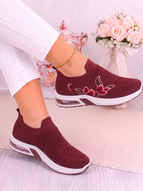 A pair of women's slip-on sneakers in burgundy with white soles and red floral embroidery on the side, shown being worn with white pants.