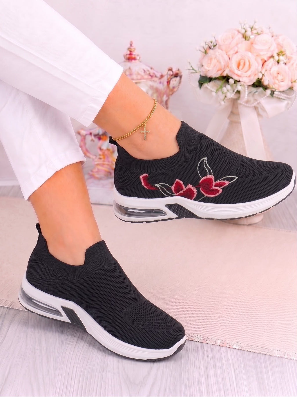 A person wearing black slip-on sneakers with red and white floral embroidery on the side, paired with white pants. The shoes feature white soles and clear air cushioning, shown against a soft pink background with decorative flowers.