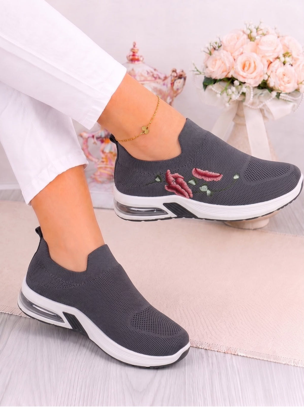 A person wearing a pair of dark gray slip-on mesh sneakers with embroidered red chili peppers and green leaves on the side, paired with white pants.