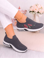 A person wearing a pair of dark gray slip-on mesh sneakers with embroidered red chili peppers and green leaves on the side, paired with white pants.