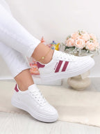 White low-top sneakers with burgundy side stripes and heel tab, shown on a model wearing white pants in a bright indoor setting.