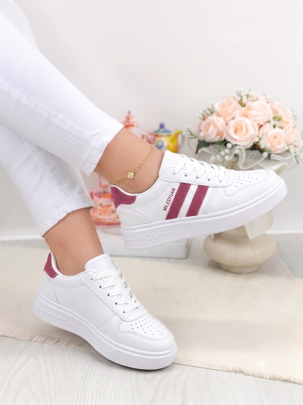 White low-top sneakers with burgundy side stripes and heel tab, shown on a model wearing white pants in a bright indoor setting.