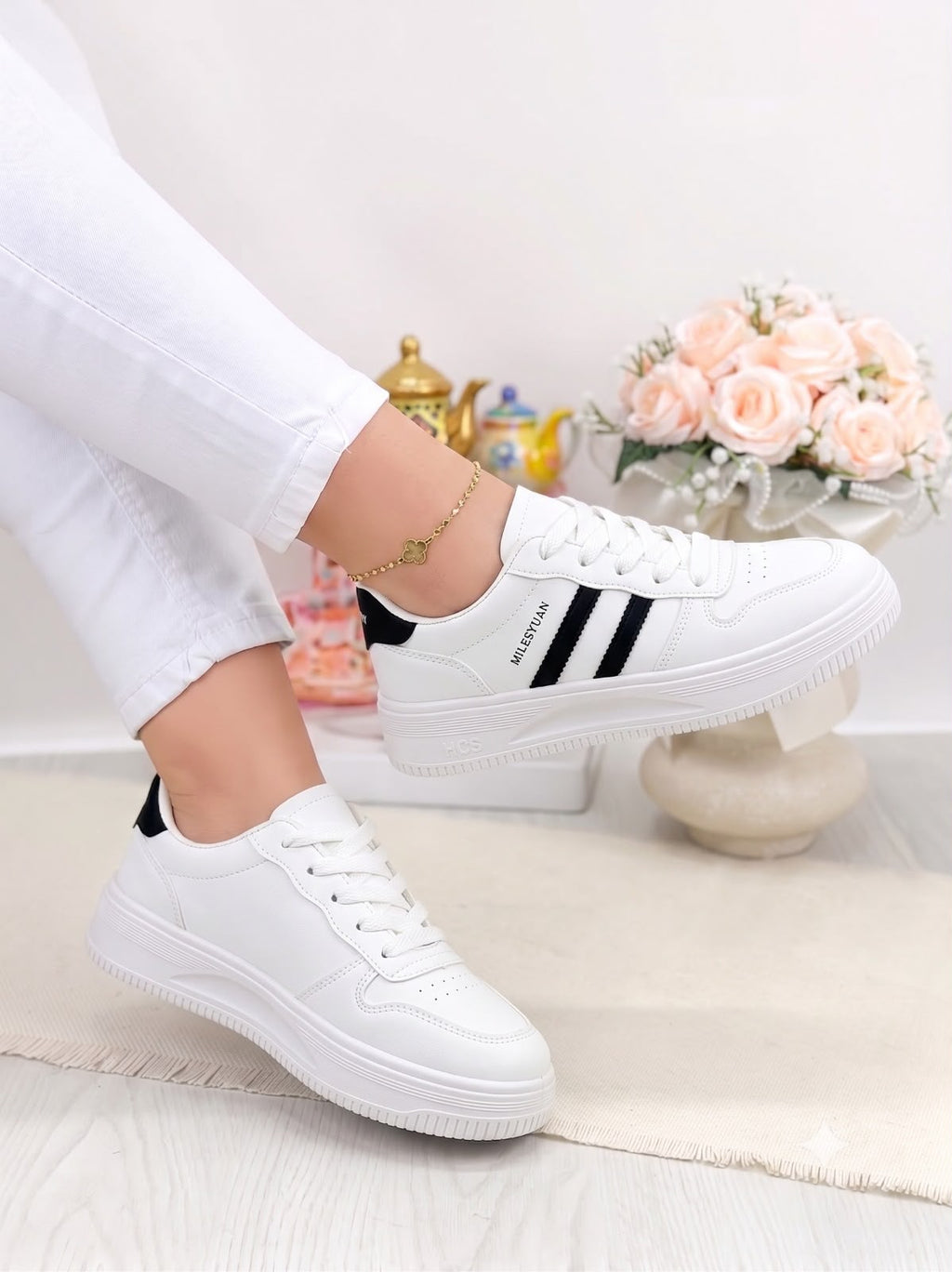 White low-top sneakers with black side stripes, shown on a model wearing white pants in a bright indoor setting with flowers in the background.