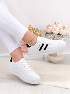 White low-top sneakers with black side stripes, shown on a model wearing white pants in a bright indoor setting with flowers in the background.