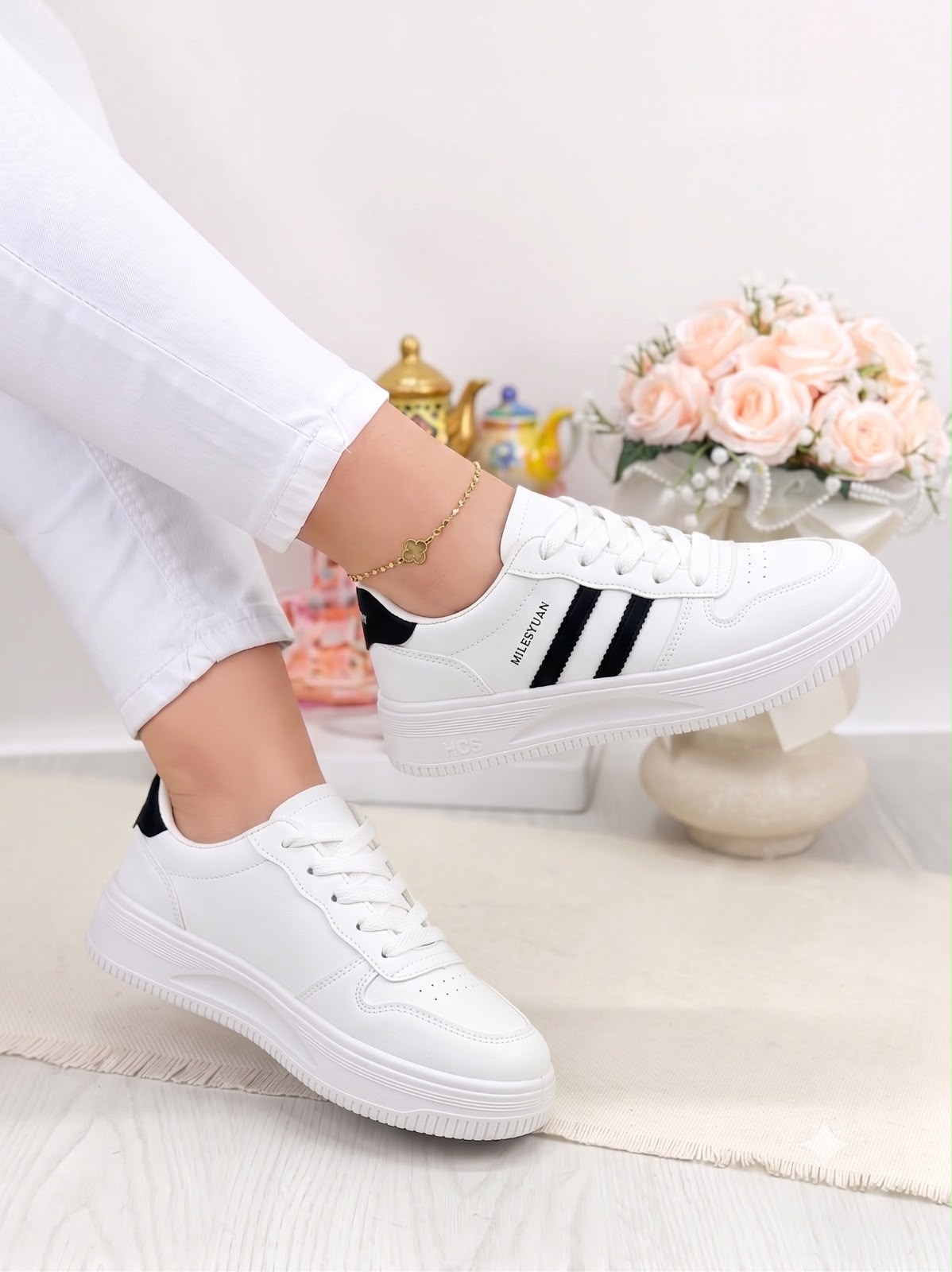 White low-top sneakers with black side stripes, shown on a model wearing white pants in a bright indoor setting with flowers in the background.