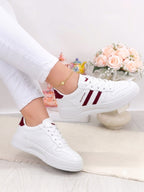 White low-top sneakers with burgundy side stripes and heel tab, shown on a model wearing white pants in a bright indoor setting with flowers in the background.