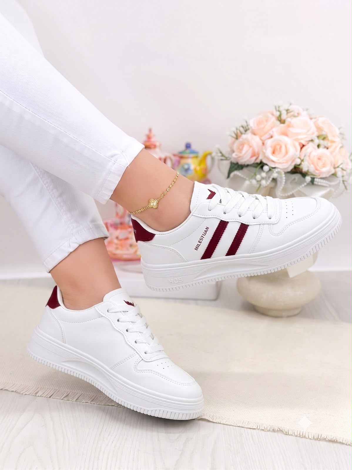 White low-top sneakers with burgundy side stripes and heel tab, shown on a model wearing white pants in a bright indoor setting with flowers in the background.