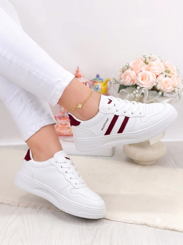 White low-top sneakers with burgundy side stripes and heel tab, shown on a model wearing white pants in a bright indoor setting with flowers in the background.
