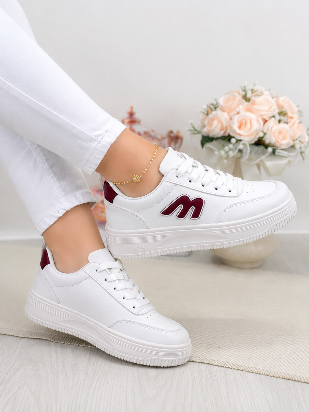 White low-top sneakers with burgundy accents, shown on a model wearing white pants. The sneakers feature a thick white platform sole and a burgundy 'm' logo on the side.