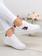 White low-top sneakers with burgundy accents, shown on a model wearing white pants. The sneakers feature a thick white platform sole and a burgundy 'm' logo on the side.