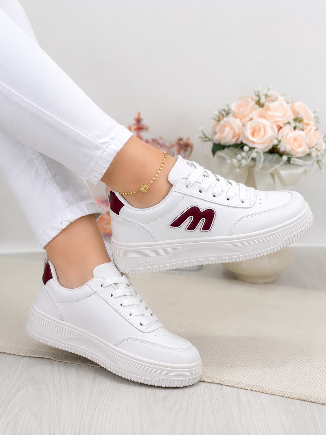 White low-top sneakers with burgundy accents, shown on a model wearing white pants. The sneakers feature a thick white platform sole and a burgundy 'm' logo on the side.