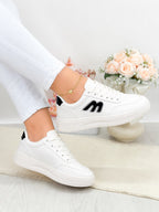 White low-top sneakers with black logo accents, shown on a model wearing white pants in a bright indoor setting with a vase of pink roses in the background.