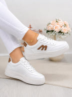 White low-top platform sneakers with a brown 'M' logo on the side, shown on a model wearing white pants. The background features a soft beige rug and a vase of pink roses.