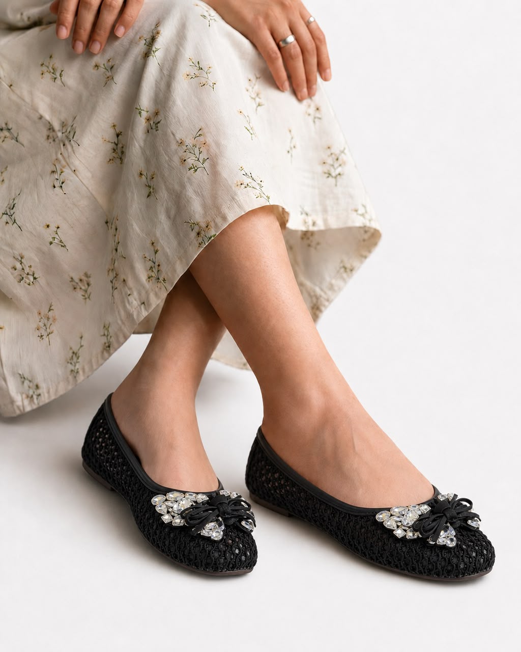 Black crocheted ballet flats with silver rhinestone bow embellishments, shown on a model wearing a cream floral maxi skirt against a light gray background.