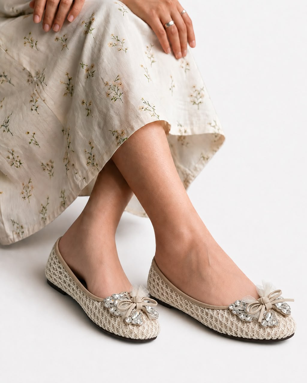 A person wearing beige woven flat shoes with decorative floral bows and rhinestone embellishments, paired with a light cream floral maxi skirt, against a clean white background.