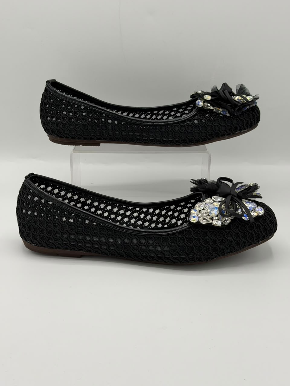 A pair of black woven flat shoes with decorative floral embellishments and rhinestone accents on the toes, displayed on a light gray background with one shoe on a clear acrylic block.